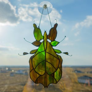 Peut inclure: Suncatcher en vitrail en forme d'insecte, aux tons verts et bruns, suspendu par une ventouse. L'insecte présente un motif détaillé en forme de feuille avec des lignes visibles. Le fond montre un ciel bleu nuageux et un paysage lointain.
