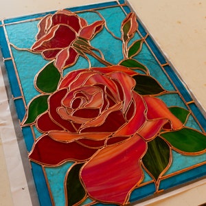 May include: A stained glass window panel featuring a red rose with green leaves on a turquoise background.