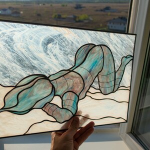 May include: A stained glass artwork depicting a figure in a blue and green pose on a sandy beach with a wave background.
