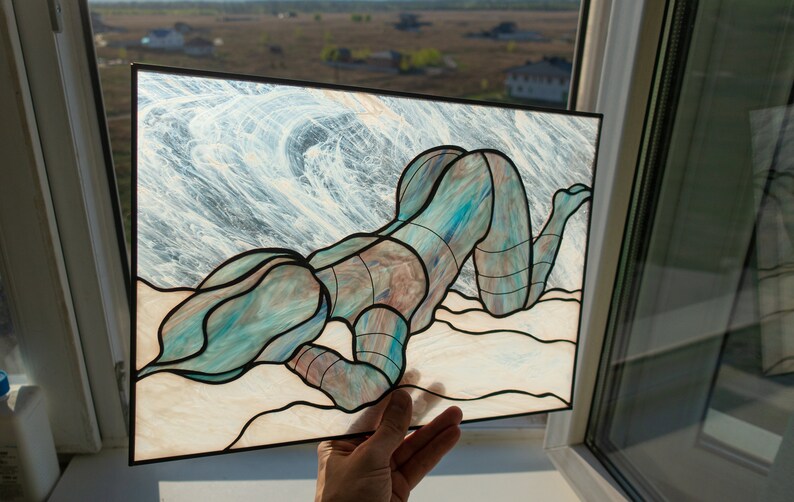 May include: A stained glass artwork depicting a figure in a blue and green pose on a sandy beach with a wave background.