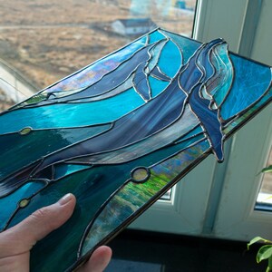 May include: Stained glass art piece featuring two whales swimming in a turquoise ocean. The whales are shades of blue and gray, with detailed fins and bodies. The glass is framed with a dark metal.