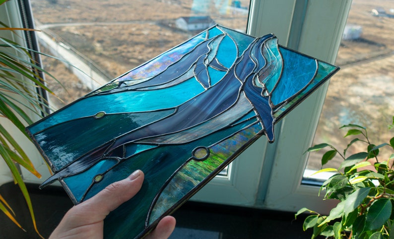 May include: Stained glass art piece featuring two whales swimming in a turquoise ocean. The whales are shades of blue and gray, with detailed fins and bodies. The glass is framed with a dark metal.