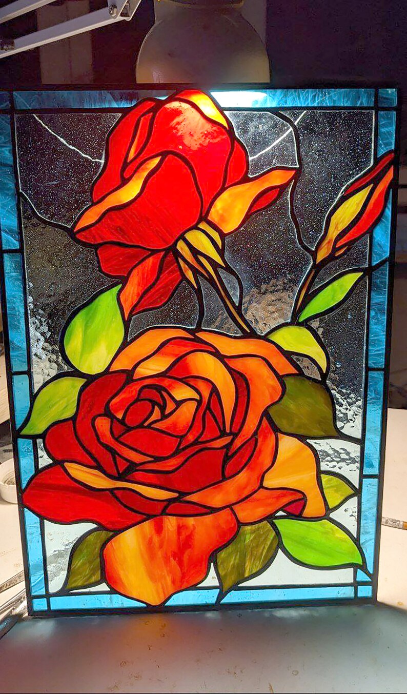 May include: A stained glass window depicting two red roses with yellow and orange accents. The roses are set against a blue background with a black and white pattern.