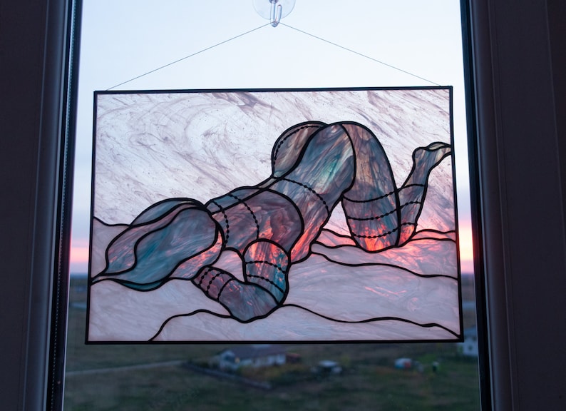 May include: A stained glass window hanging in a window frame. The stained glass depicts a person in a kneeling position with their head resting on their hand. The stained glass is made of various shades of blue, green, red, and orange glass.