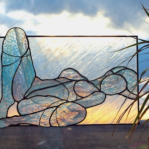 May include: A stained glass window depicting a stylized figure in a reclining pose. The figure is made of blue and green glass pieces, with a textured, iridescent finish. The background is a light blue and white, with a hint of yellow at the bottom.