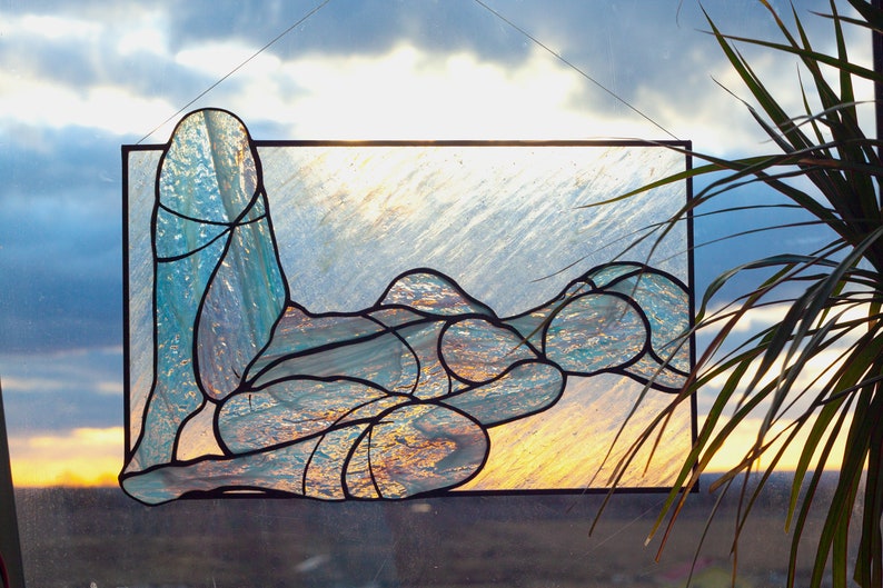 May include: A stained glass window depicting a stylized figure in a reclining pose. The figure is made of blue and green glass pieces, with a textured, iridescent finish. The background is a light blue and white, with a hint of yellow at the bottom.