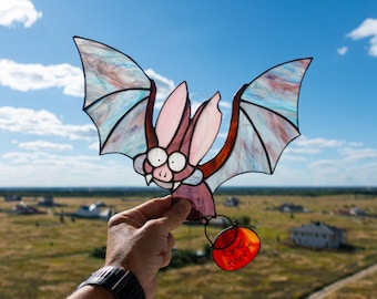 May include: A stained glass bat with a pink body and blue and purple wings. The bat is holding a red pumpkin with a scary face.
