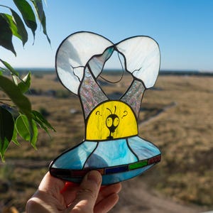 May include: A stained glass UFO with a blue and yellow design. The UFO has a small alien figure inside with question marks above its head.