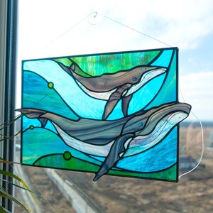 May include: Stained glass panel featuring two whales swimming in a turquoise and green ocean. The whales are shades of gray and brown, with white accents. The panel is rectangular and hangs in front of a window.