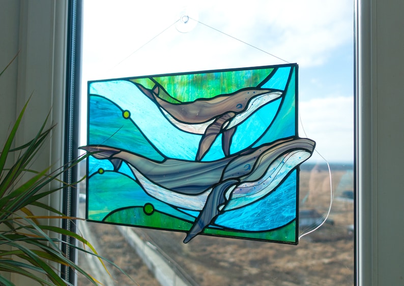 May include: Stained glass panel featuring two whales swimming in a turquoise and green ocean. The whales are shades of gray and brown, with white accents. The panel is rectangular and hangs in front of a window.