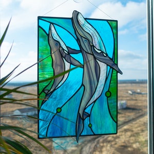May include: Stained glass panel featuring two whales swimming in blue and green water. The whales are depicted in shades of gray and white, with detailed fins and bodies. The panel is rectangular and hangs in front of a window.