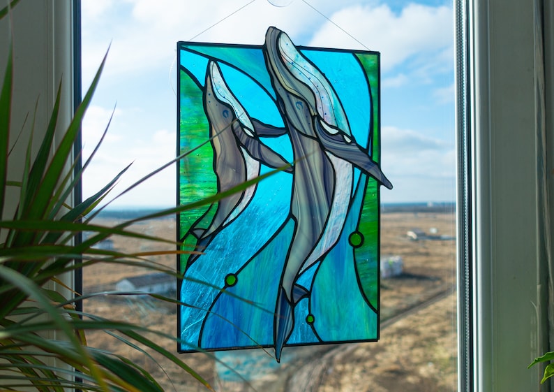 May include: Stained glass panel featuring two whales swimming in blue and green water. The whales are depicted in shades of gray and white, with detailed fins and bodies. The panel is rectangular and hangs in front of a window.