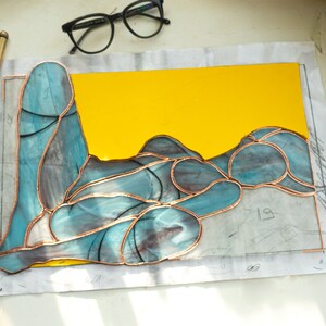 May include: A stained glass piece depicting a reclining figure with a yellow background. The glass is cut into various shapes and joined together with copper foil.