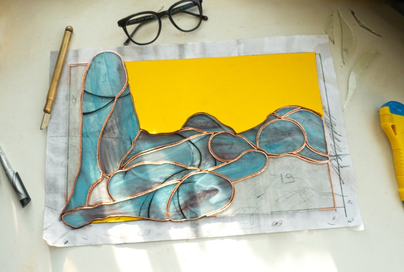 May include: A stained glass piece depicting a reclining figure with a yellow background. The glass is cut into various shapes and joined together with copper foil.