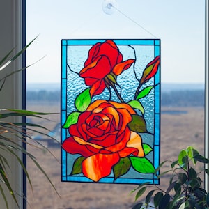 May include: A stained glass window hanging in a window frame, depicting three red roses with green leaves and stems on a blue background.