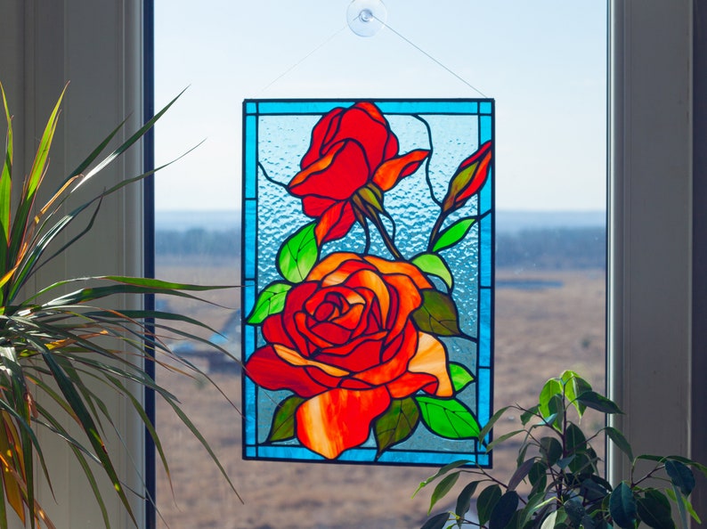 May include: A stained glass window hanging in a window frame, depicting three red roses with green leaves and stems on a blue background.