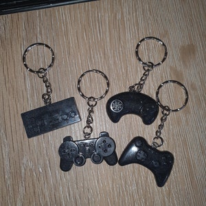 May include: Four black keychains with silver keyrings. The keychains are shaped like retro video game controllers and a cassette tape. The keychains are on a light brown wooden surface.