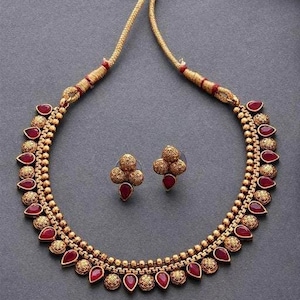 Op de afbeelding: Een gouden ketting met rode edelsteenaccenten en bijpassende oorbellen. De ketting heeft een delicate ketting met kleine ronde kralen en hangende traanvormige stenen. De oorbellen zijn klein en hebben drie ronde stenen.