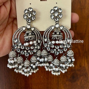 May include: A pair of silver earrings with a round design and multiple layers of dangling pearls. The earrings feature a large, round, clear stone in the center and smaller clear stones around the edges. The earrings are labeled "B TRENDY EXCLUSIVE JEWELLERY".