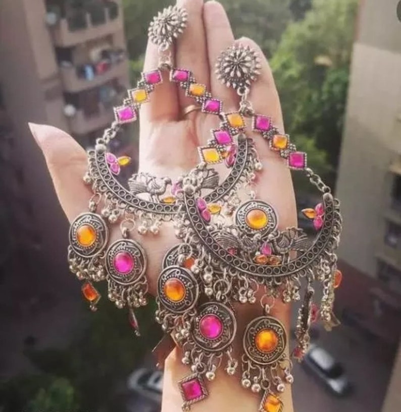 May include: A pair of large silver earrings with pink and orange glass stones. The earrings have a crescent moon shape with intricate details and dangling charms.