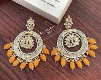 May include: A pair of gold earrings with a round design. The earrings have a white and gold center with a floral pattern. The earrings are adorned with small white beads and orange beads.