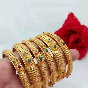 Op de afbeelding: Een set van zes gouden armbanden met ingewikkelde ontwerpen en groene en rode edelstenen. De armbanden zijn op elkaar gestapeld.