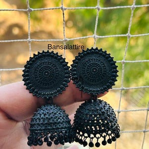 May include: A pair of black jhumka earrings with a round, ornate design. The earrings have a dangling, bell-shaped bottom with small beads.
