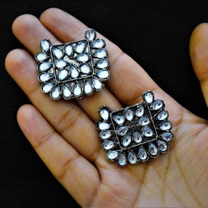 May include: A pair of silver earrings with clear stones arranged in a square shape. The earrings are on a hand.