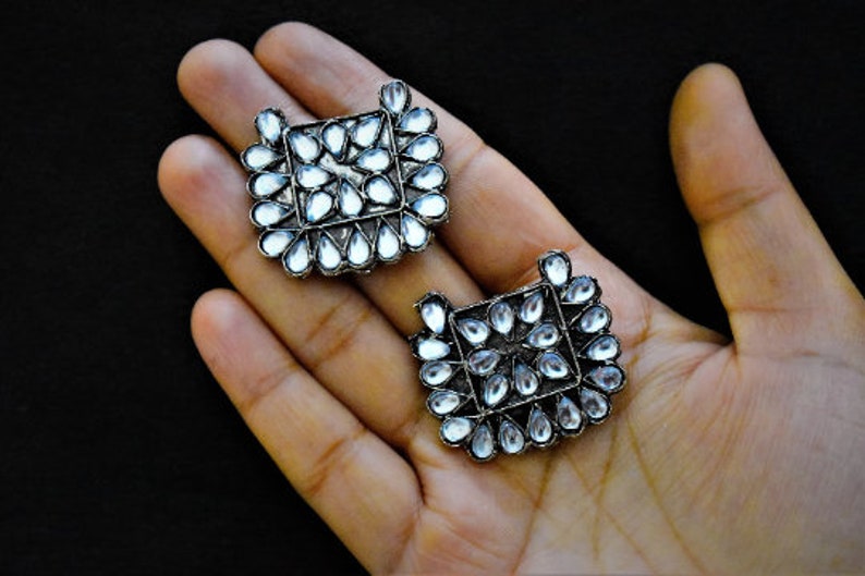 May include: A pair of silver earrings with clear stones arranged in a square shape. The earrings are on a hand.