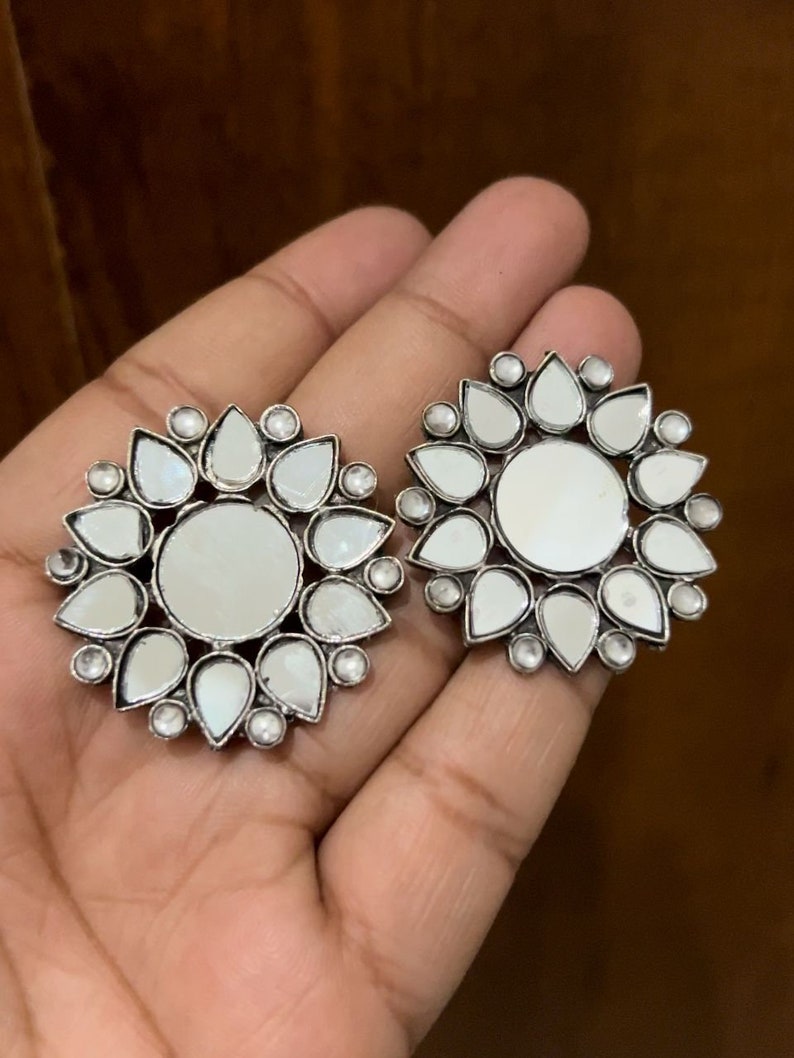 May include: A pair of silver tone earrings with a circular design. Each earring features a central mirror surrounded by smaller mirrored teardrop shapes.