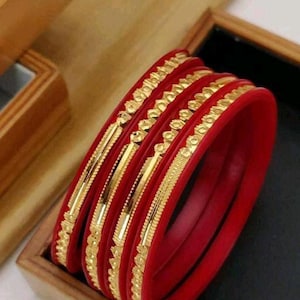 Op de afbeelding: Set van vier gouden armbanden met ingewikkelde ontwerpen, gelaagd op rode armbanden.