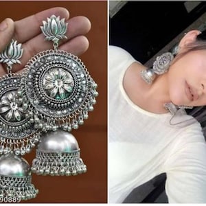 May include: A pair of silver dangle earrings with a floral design and a bell-shaped bottom. The earrings are made of metal and have a traditional Indian style.