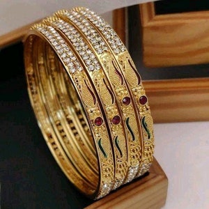 May include: A set of four gold bangles with intricate designs and red and green accents. The bangles are adorned with sparkling white stones.