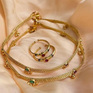 May include: A set of three gold anklets with green and red gemstone accents. The anklets are made of delicate, interwoven chains.