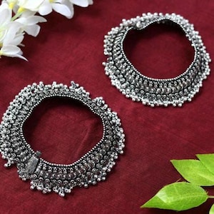 May include: A pair of silver anklets with intricate designs and dangling beads. The anklets are on a red background.