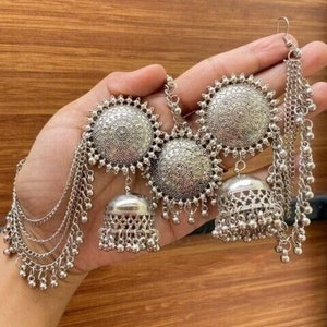 May include: A pair of silver dangle earrings with multiple chains and decorative bells. The earrings feature a large round disc with intricate designs.