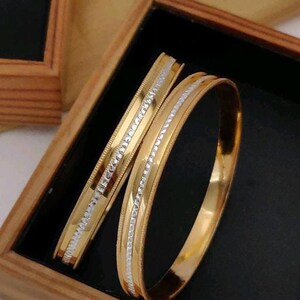 May include: Two gold bangles with a white metal design. The bangles are stacked on top of each other and are in a wooden box.
