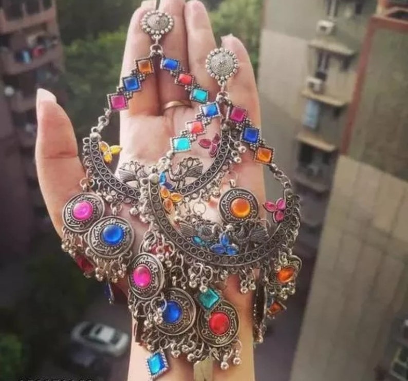 May include: A pair of silver earrings with colorful glass stones and intricate metalwork. The earrings feature a crescent moon shape with dangling charms and a long, beaded drop.