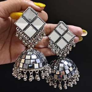 Peut inclure: Une paire de boucles d'oreilles en argent avec un motif miroir. Les boucles d'oreilles présentent un dessus carré avec quatre carrés miroir et un dessous rond avec une mosaïque de carrés miroir. Les boucles d'oreilles sont suspendues à l'oreille d'une personne.