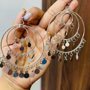 May include: Silver hoop earrings with three tiers of dangling silver coins. The coins are a mix of silver, gold, and blue.