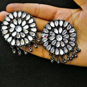 May include: A pair of silver earrings with a round, floral design. The earrings have a black oxidized finish and are adorned with clear, teardrop-shaped stones.