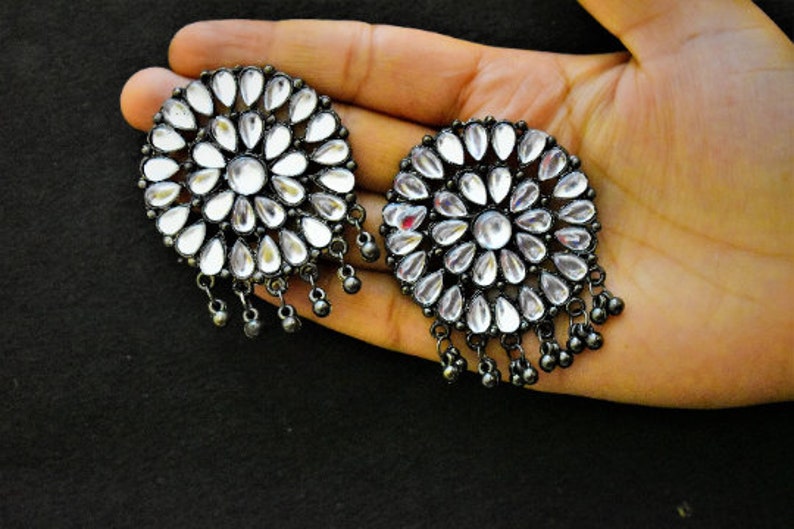 May include: A pair of silver earrings with a round, floral design. The earrings have a black oxidized finish and are adorned with clear, teardrop-shaped stones.