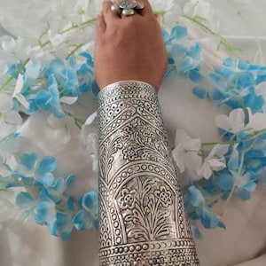 Op de afbeelding: Een zilveren geoxideerde metalen armband met een ingewikkeld bloemenpatroon. De armband is breed en bedekt het grootste deel van de onderarm.