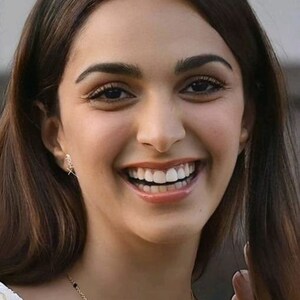 May include: A woman with long dark brown hair is smiling. She is wearing a white and yellow top with a gold chain necklace with black beads. She has a red dot on her forehead.