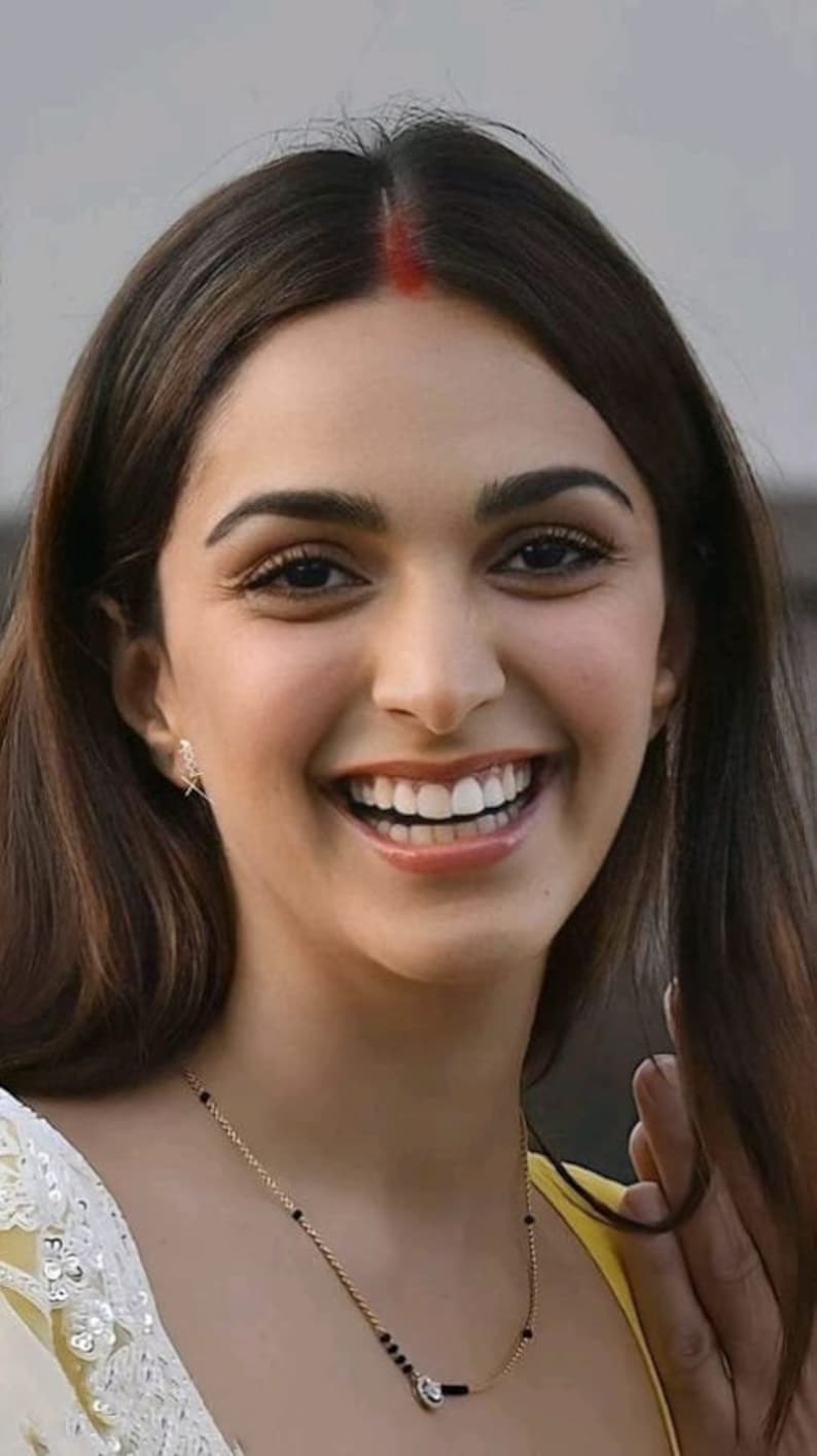 May include: A woman with long dark brown hair is smiling. She is wearing a white and yellow top with a gold chain necklace with black beads. She has a red dot on her forehead.