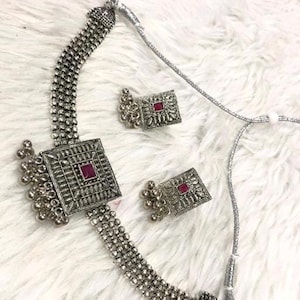 May include: A silver necklace with a square pendant featuring a pink stone. The necklace has a chain made of small silver beads. The pendant is attached to a silver chain with a tassel. The necklace comes with a pair of matching earrings.