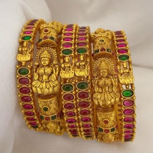 May include: Gold-toned bangles with intricate detailing and embedded gemstones. The bangles feature a central figure and are adorned with red and green stones. The jewelry is displayed on a white surface, showcasing the ornate design.