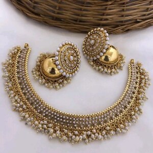 Könnte beinhalten: Ein goldenes Halsketten- und Ohrring-Set mit weißen Perlenakzenten. Die Halskette hat eine Halbmondform mit einer zarten Kette und Perlentropfen. Die Ohrringe sind rund mit einem ähnlichen Design.