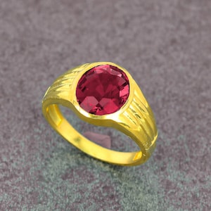 May include: A gold ring featuring a large, round, faceted, deep red gemstone. The ring has a textured, ridged band and a bright, polished finish. The gemstone is the focal point, set in a simple, elegant design.