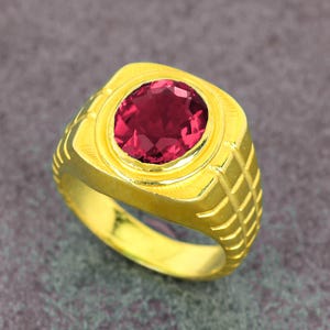 May include: A gold-colored ring with a large, oval-shaped, deep red gemstone. The ring has a textured band with a geometric pattern and a polished finish, showcasing the gemstone's brilliance.